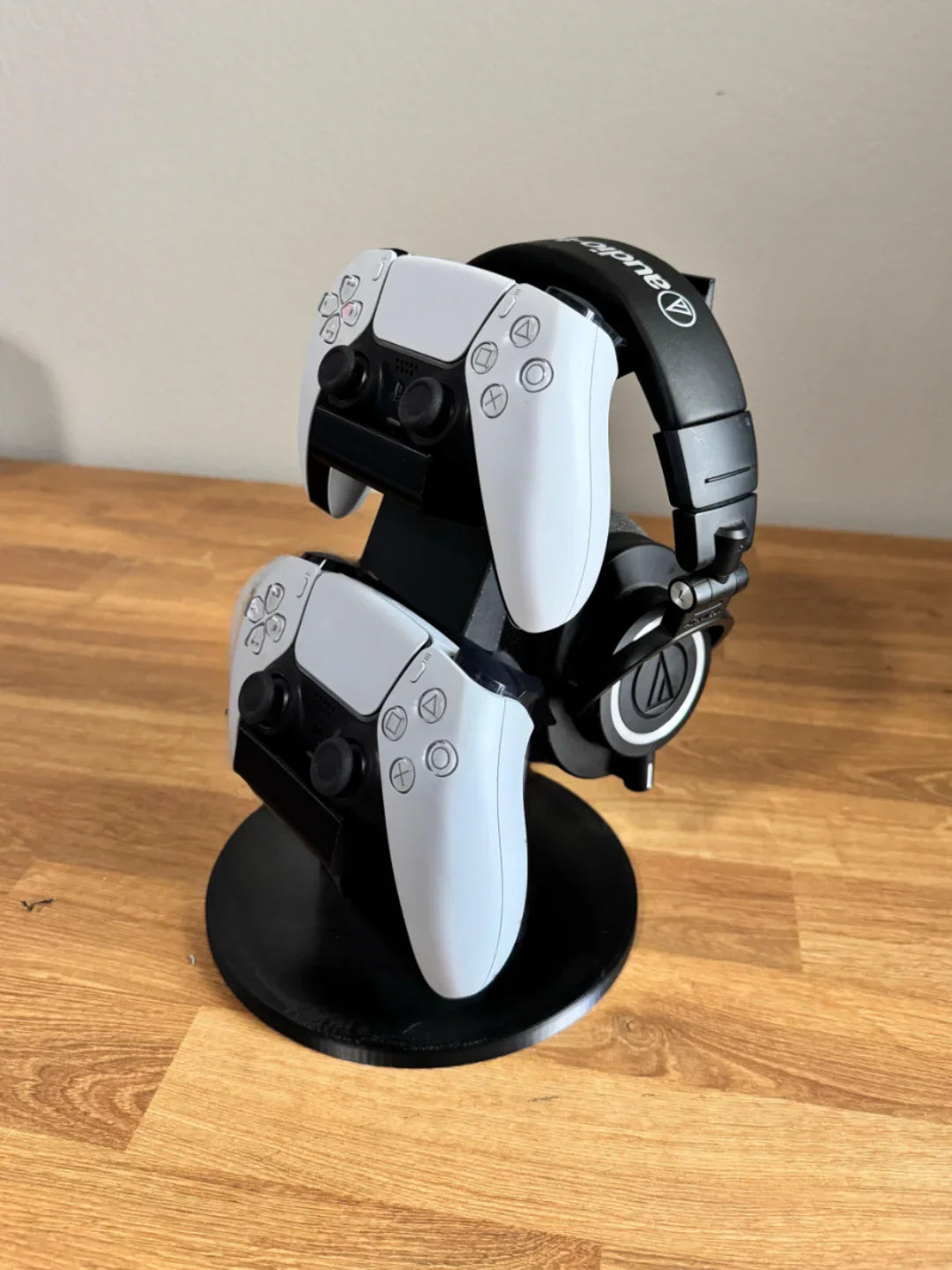3D printed black stand holding two PlayStation controllers and headphones on wood surface