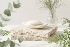 White soap dish with a bar of soap styled on stone blocks with soft greenery.
