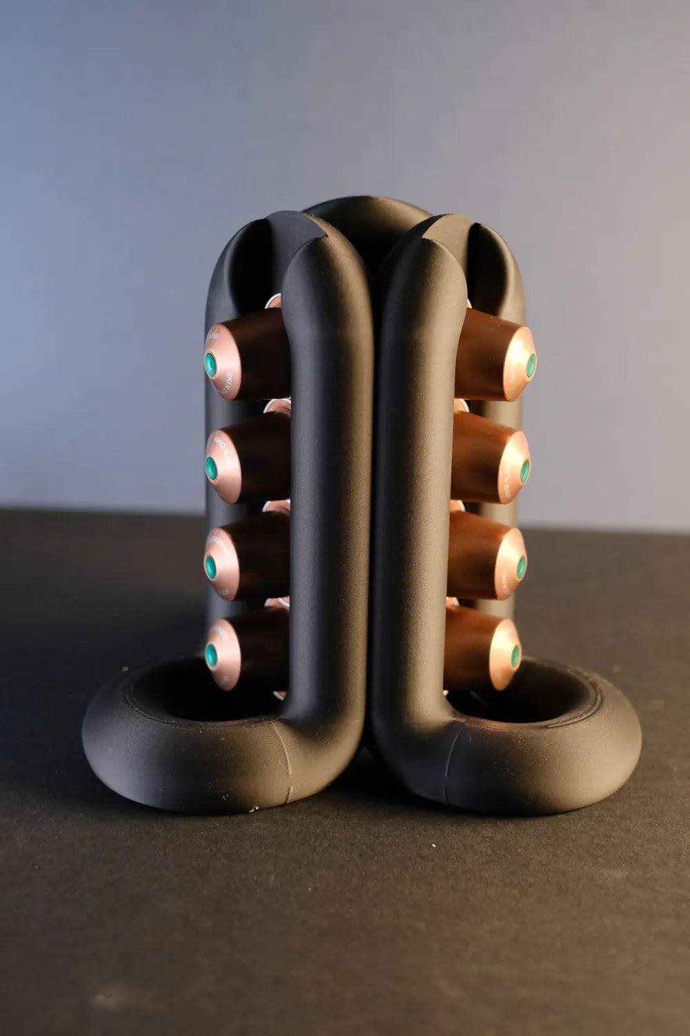 Black three-way coffee pod holder standing on a dark surface with capsules arranged on three sides
