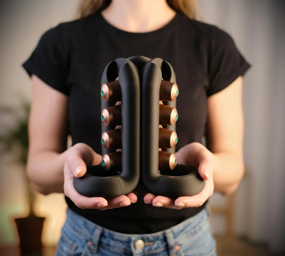 Black sculptural coffee pod holder held in both hands with capsules visible on opposing sides